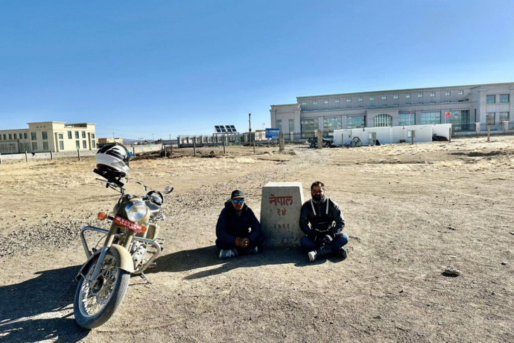 Two bikers sitting beside a Nepal border marker with a Royal Enfield bike and buildings in the background.
