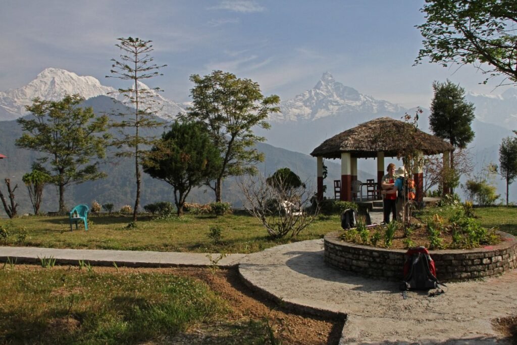 "Tourists relax in a garden with a thatched gazebo in Dhampus, Nepal, with Machapuchare and Annapurna peaks in the background."