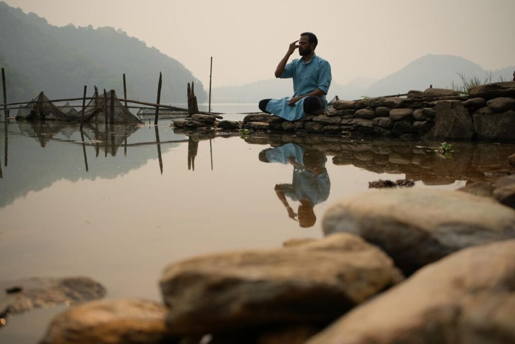 "Man practicing pranayama breathing technique by a calm riverside with reflections in the water and misty hills in the background"