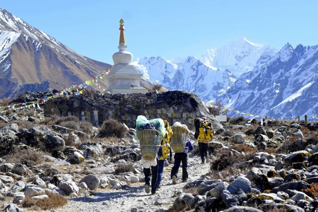 "Porters walking on a rocky Langtang trail towards a white stupa, with snow-covered peaks in the background."